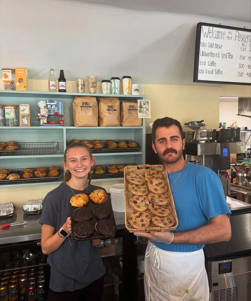 Two staff behind the counter holding trays of muffins and cookies, with the chalkboard drinks menu behind them