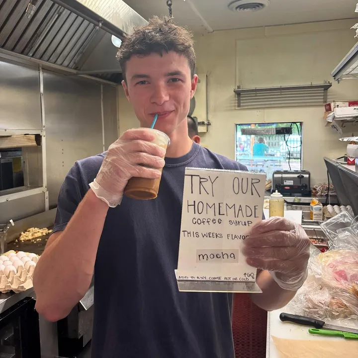 A staff member with an iced coffee beside a handwritten homemade coffee syrup sign